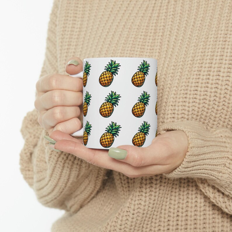 A person is holding a white mug with a repeating pattern of pineapples printed on it.