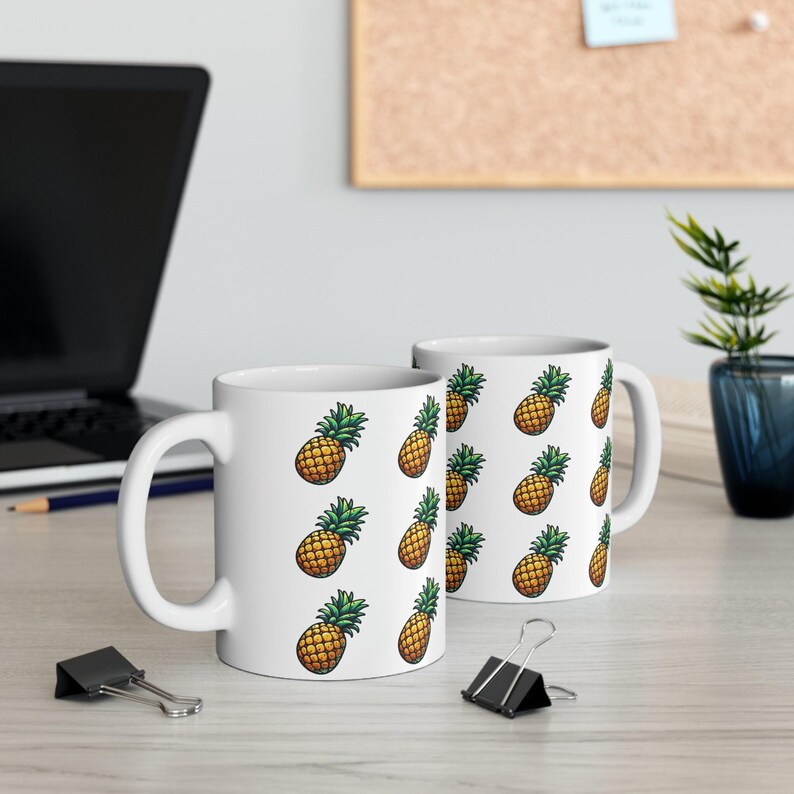two white mugs with pineapple designs on them, placed on a desk with a laptop, a plant, and some office supplies.
