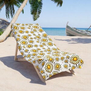 A beach chair with a white and yellow floral patterned blanket is placed on a sandy beach, with palm trees and a boat visible in the background.