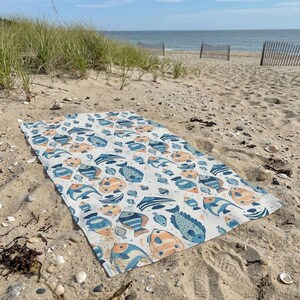 A colorful beach towel with a fish pattern lies on the sandy beach, with the ocean and a wooden fence visible in the background.