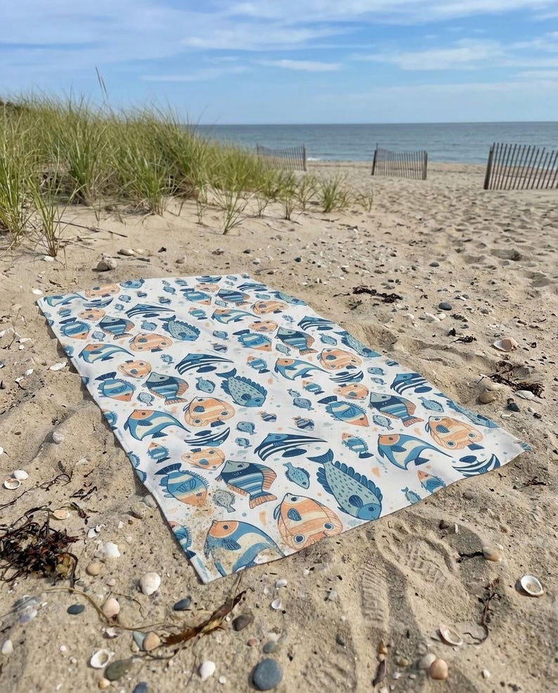 A colorful beach towel with a fish pattern lies on the sandy beach, with the ocean and a wooden fence visible in the background.