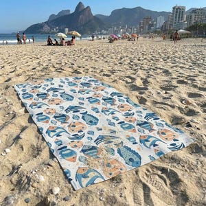 a beach scene with a colorful towel spread out on the sand, featuring a pattern of fish and other marine life. In the background, there are people enjoying the beach and the ocean, with buildings visible in the distance.
