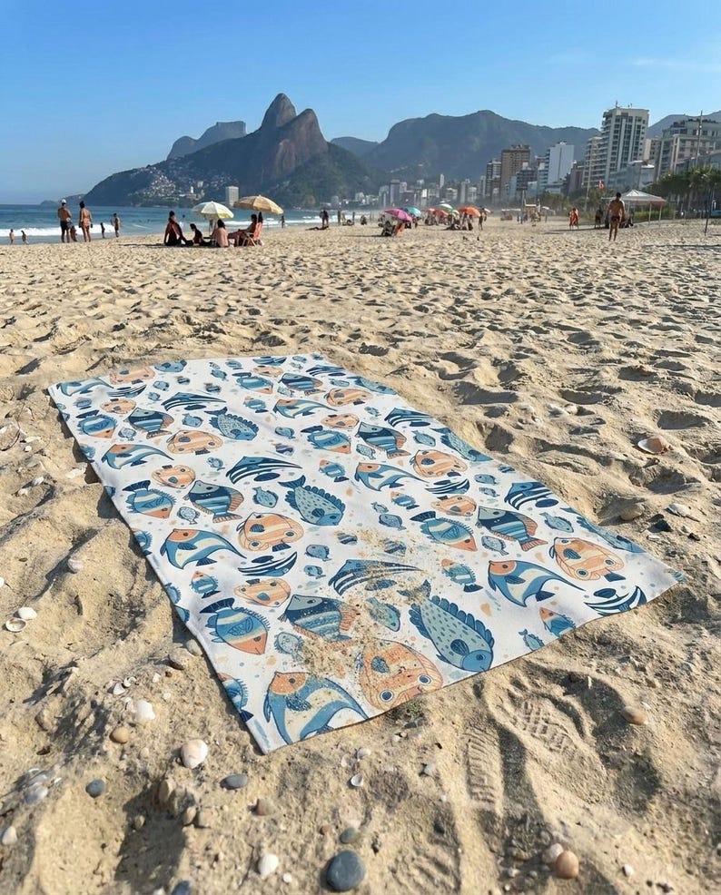 a beach scene with a colorful towel spread out on the sand, featuring a pattern of fish and other marine life. In the background, there are people enjoying the beach and the ocean, with buildings visible in the distance.