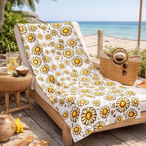 A comfortable lounge chair with a yellow and white floral patterned blanket, placed on a wooden deck overlooking a beach. The chair is accompanied by a straw umbrella, a woven basket, and a pair of sandals.
