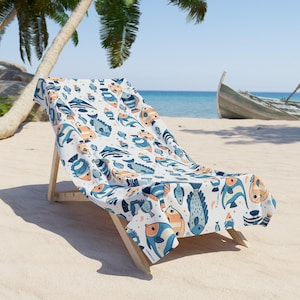 A colorful beach towel with fish patterns is draped over a wooden chair on a sandy beach, with palm trees and a boat visible in the background.