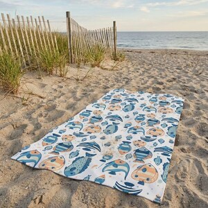 A colorful beach towel with a fish pattern lies on the sandy beach, with a wooden fence and grassy area in the background.