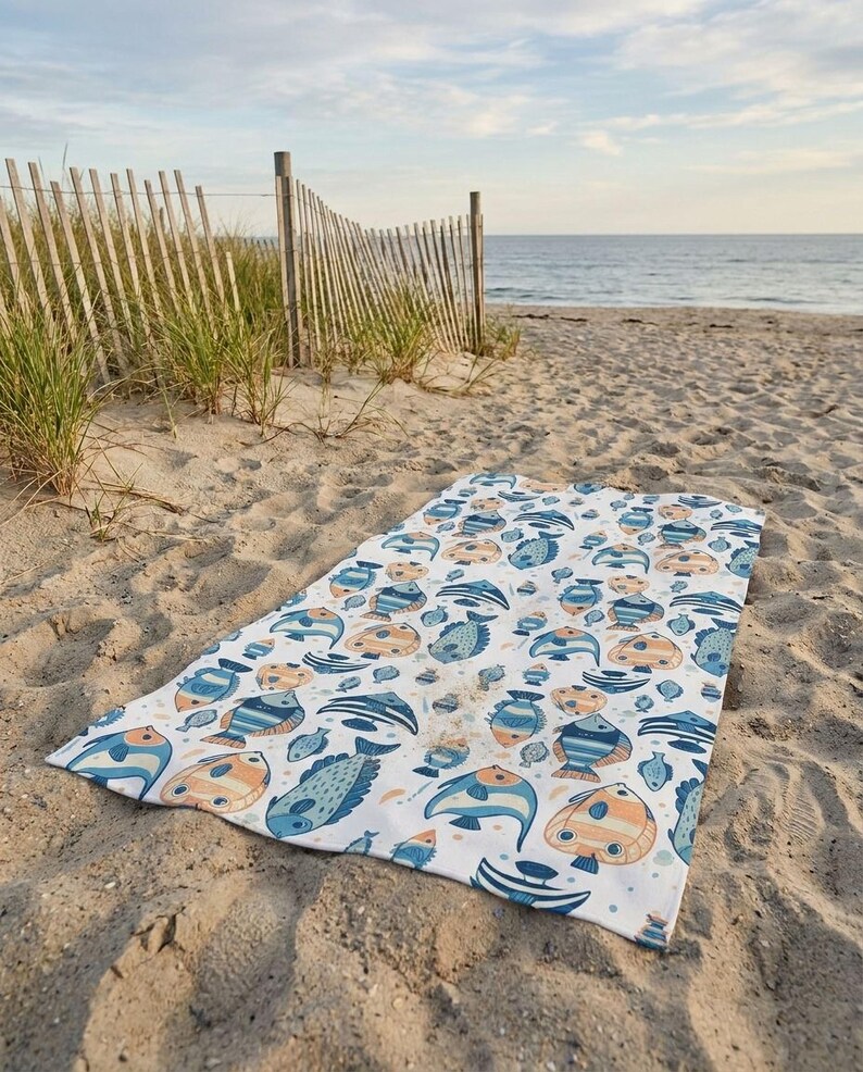 A colorful beach towel with a fish pattern lies on the sandy beach, with a wooden fence and grassy area in the background.