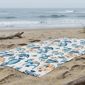 A colorful beach towel with a fish pattern lies on the sandy beach, with the ocean and waves visible in the background.