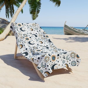 A beach chair with a patterned towel draped over it, situated on a sandy beach with palm trees and a boat visible in the background.
