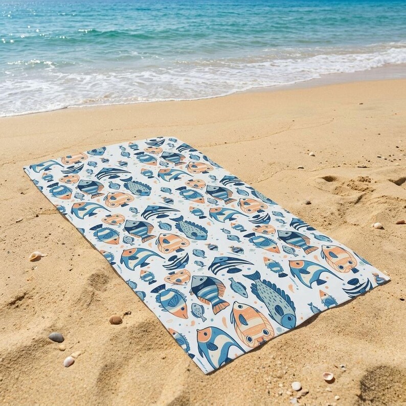 A colorful beach towel with a fish pattern lies on the sandy beach, with the ocean visible in the background.