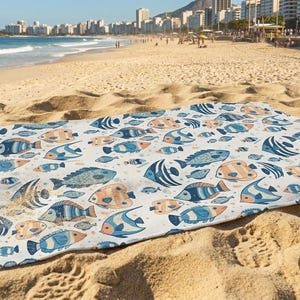 A colorful beach towel with a fish pattern is spread out on the sandy beach, with the ocean and a city skyline visible in the background.