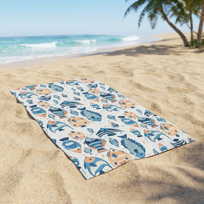 A colorful beach towel with a fish pattern is spread out on the sandy beach, with the ocean and palm trees visible in the background.