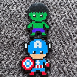 Puede incluir: Dos figuras de pixel art: Hulk, verde con pelo negro y pantalones cortos morados, y Capitán América, con traje azul con estrella blanca, sosteniendo un escudo. Las figuras están hechas de pequeñas cuentas de colores.