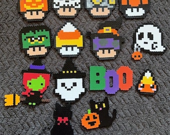 May include: A collection of Halloween-themed pixel art decorations. The designs include pumpkins, skulls, mushrooms, ghosts, witches, candy corn, and black cats. The word "BOO" is also spelled out in colorful letters. These are likely made from fuse beads.