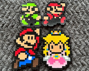 May include: Four pixel art figures of characters from the Super Mario Bros. video game series. The characters include Mario, Luigi, and Princess Peach. The figures are made of colorful plastic beads.