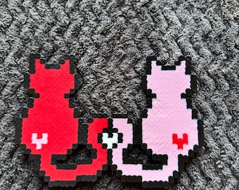 Può includere: Due spille a forma di gatto in pixel art. Una è rossa con un design bianco e nero, l'altra è rosa con un design bianco e rosso. Entrambe hanno contorni neri e sono su uno sfondo grigio strutturato.