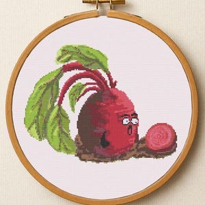 Puede incluir: Bordado de punto de cruz de una remolacha de dibujos animados con hojas verdes y una expresión de sorpresa. La remolacha es roja con una base marrón, en un bastidor de bordado de madera.