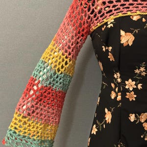 Könnte beinhalten: Ein langärmeliger Häkel-Bolero mit einem mehrfarbigen Netzmuster in Koralle, Gelb und hellem Türkis, getragen über einem ärmellosen schwarzen Kleid mit einem Blumenmuster aus hellorangen Blüten und grünen Blättern.