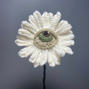 May include: A crocheted white daisy with a green and brown eye in the center. The daisy is on a black stem.