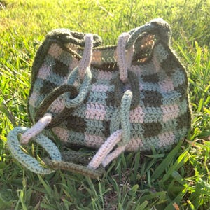 May include: A green and white crocheted tote bag with a checkered pattern. The bag has long straps that are tied together at the top.