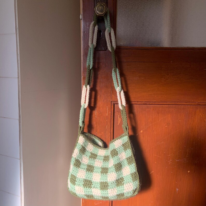 May include: A green and white checkered crocheted bag with a long chain strap made of beige and green yarn.