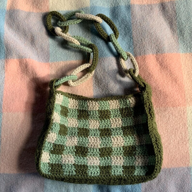 May include: A green and white crocheted checkered bag with a thick chain strap. The bag is made of yarn and has a rectangular shape.