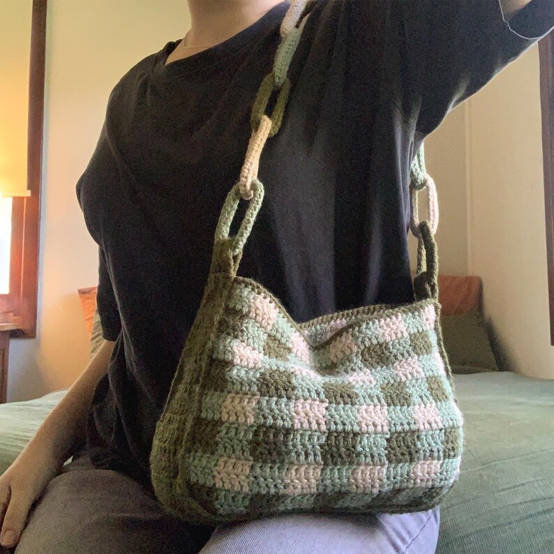 May include: A green and white crocheted shoulder bag with a checkered pattern. The bag has a long strap and is worn over the shoulder.