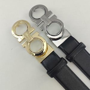 May include: Two black belts with textured leather straps and metal Gancini buckles. One buckle is polished gold-tone metal, and the other is polished silver-tone metal. Each buckle features the engraved Salvatore Ferragamo brand name.