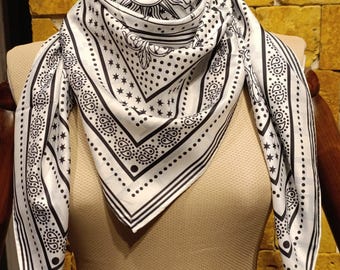 May include: A white and black patterned scarf with a sun and star design. The scarf is draped over a mannequin, showcasing its square shape and intricate details. The design includes paisley and dotted borders.