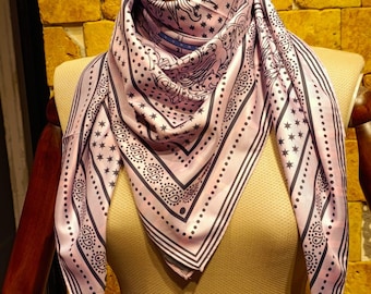 May include: A light pink silk scarf with a black and white star and paisley pattern. The scarf is draped over a mannequin, showcasing its square shape and intricate design. The scarf has a border with stripes and dots.