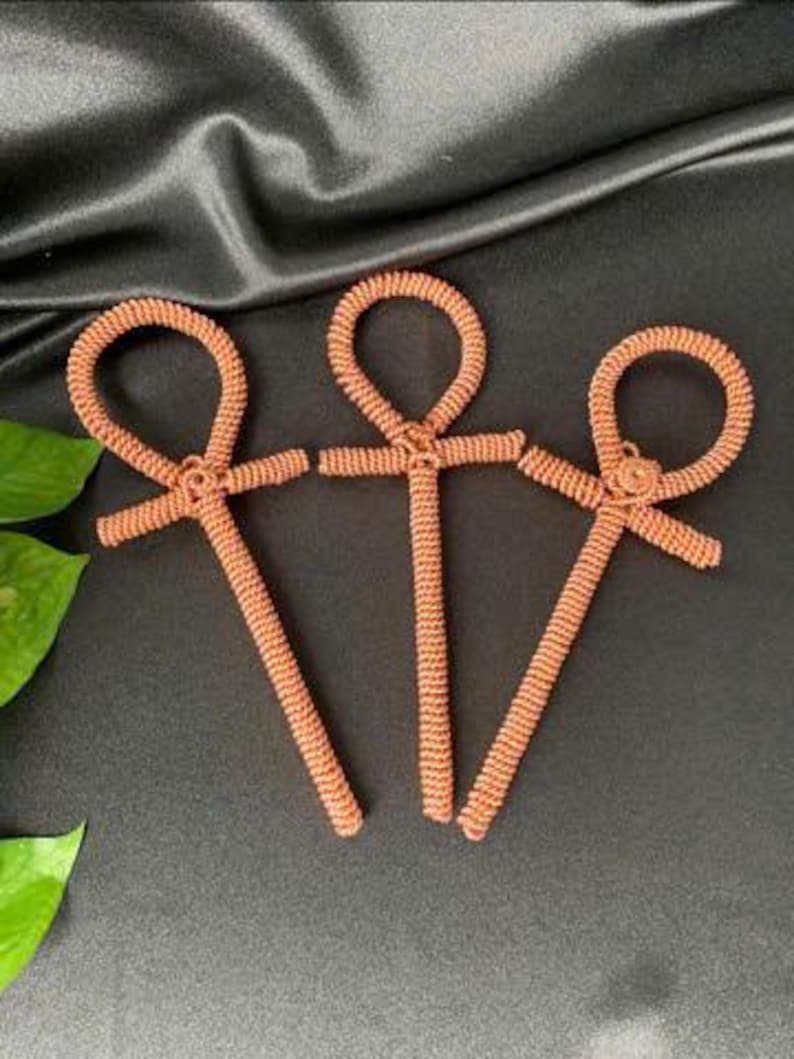 May include: Three copper-colored ankh symbols, each with a looped top and a cross-like shape below. The ankhs are made of tightly wound wire, and are arranged on a dark, textured surface. The ankhs are a symbol of life.