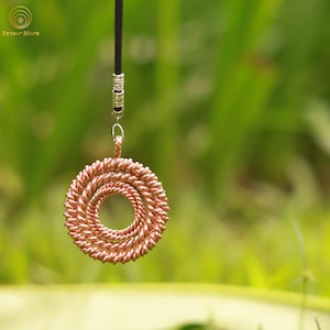 May include: A copper-colored pendant necklace with a spiral design. The pendant is suspended from a black cord with silver-colored metal accents. The background is a blurred green.