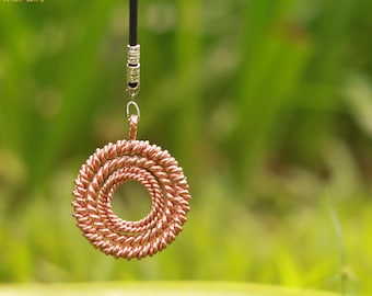 May include: A copper-colored pendant necklace with a spiral design. The pendant is suspended from a black cord with silver-colored metal accents. The background is a blurred green.