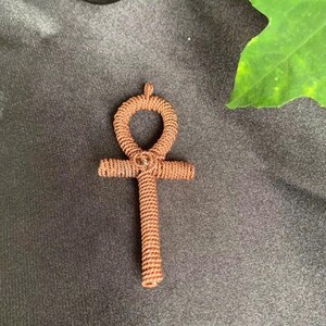 May include: A brown, woven ankh pendant. The ankh, a symbol of life, features a looped top, a horizontal bar, and a vertical stem. The pendant is made of tightly woven brown material, and is set against a dark background.