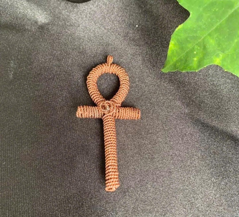 May include: A brown, woven ankh pendant. The ankh, a symbol of life, features a looped top, a horizontal bar, and a vertical stem. The pendant is made of tightly woven brown material, and is set against a dark background.