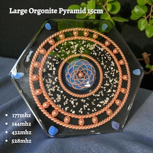 Op de afbeelding: Een grote, zeshoekige orgonietpiramide, ongeveer 15 cm breed, met koperdraadspiralen en een centraal blauw ontwerp. De piramide heeft een zwarte basis met zilveren spikkels en blauwe accenten. De tekst luidt "Large Orgonite Pyramid 15cm" en vermeldt frequenties.