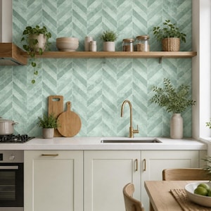 May include: A kitchen featuring light green herringbone tiles on the wall, a wooden shelf holding ceramic bowls and glass jars, a stainless steel tap over a sink, light-coloured cabinets, and wooden cutting boards on the white worktop.