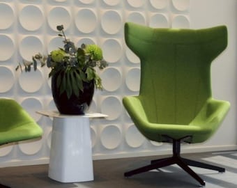May include: A modern interior scene featuring a high-backed, lime green armchair with a dark base. A white side table holds a dark vase with green foliage. The background has a textured white wall with circular patterns.