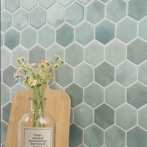 Może przedstawiać: Zbliżenie ściany z sześciokątnymi płytkami w odcieniach niebieskiego i zielonego. Przezroczysty szklany wazon z etykietą i bukietem białych kwiatów stoi przed drewnianą deską do krojenia. Na pierwszym planie rogalik.