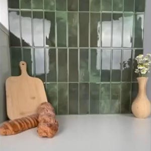 Puede incluir: Una tabla de cortar de madera, una barra de pan y un jarrón decorativo con flores están dispuestos sobre una superficie blanca. El fondo presenta azulejos rectangulares de color verde oscuro. La escena evoca una cocina o comedor.