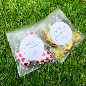 May include: Two clear plastic bags with white polka dots. One bag contains pink heart-shaped chocolates with a white circle sticker that says "HER favourite" with a small heart. The other bag contains gold-wrapped chocolates with a white circle sticker that says "HIS favourite" with a small heart.