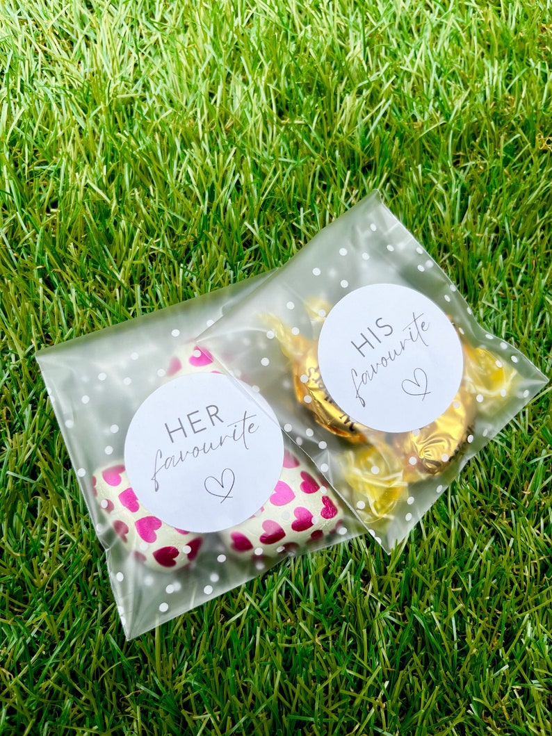 May include: Two clear plastic bags with white polka dots. One bag contains pink heart-shaped chocolates with a white circle sticker that says "HER favourite" with a small heart. The other bag contains gold-wrapped chocolates with a white circle sticker that says "HIS favourite" with a small heart.