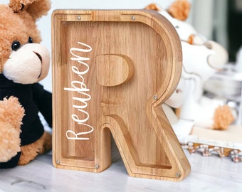 May include: Wooden piggy bank shaped like the letter R with the name Reuben engraved on the front. The piggy bank is light brown and has a slot on the top for coins.