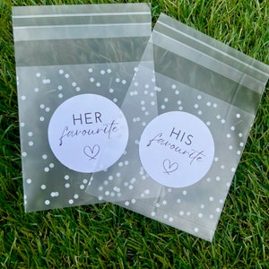 May include: Two clear plastic bags with white polka dots and a white circle with black text that says "HER favourite" and "HIS favourite" with a heart symbol.