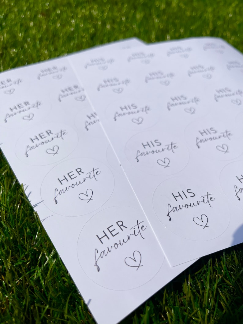 May include: A sheet of white stickers with black text and a heart design. The stickers say "HER favourite" and "HIS favourite".