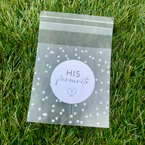 May include: A clear plastic bag with a white polka dot pattern. The bag has a sticker on it that says "HIS favourite" with a heart symbol.