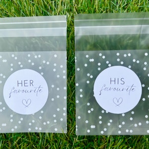 May include: Two clear plastic bags with white polka dots and a white circle sticker on each. The sticker on the left bag says "HER favourite" with a heart symbol. The sticker on the right bag says "HIS favourite" with a heart symbol.