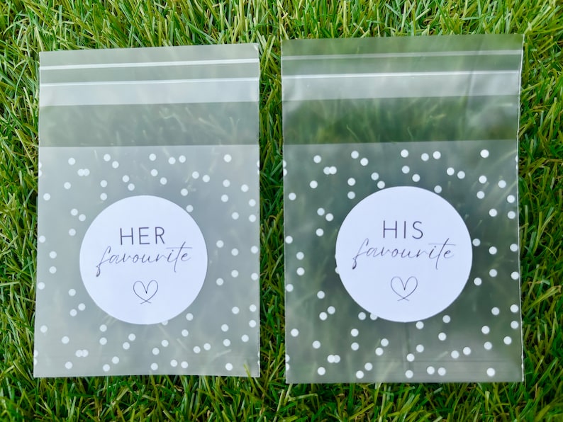 May include: Two clear plastic bags with white polka dots and a white circle sticker on each. The sticker on the left bag says "HER favourite" with a heart symbol. The sticker on the right bag says "HIS favourite" with a heart symbol.