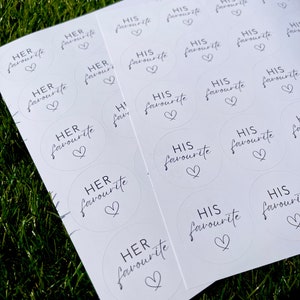 May include: A sheet of white paper with round stickers printed with the text "His favourite" and "Her favourite" in black script with a heart symbol.
