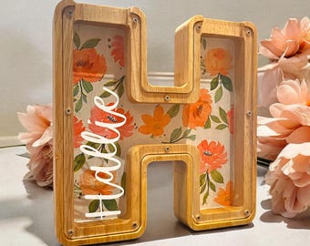 May include: A wooden letter H with a floral pattern and a clear acrylic insert. The letter is decorated with a white vinyl name "Hollie".